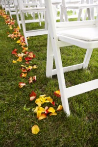 White wedding chairs on a green lawn with flower petals sprinkled down the isle.
