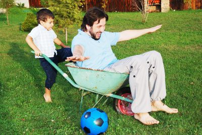 Kid pushing dad in wheelbarrow on green grass